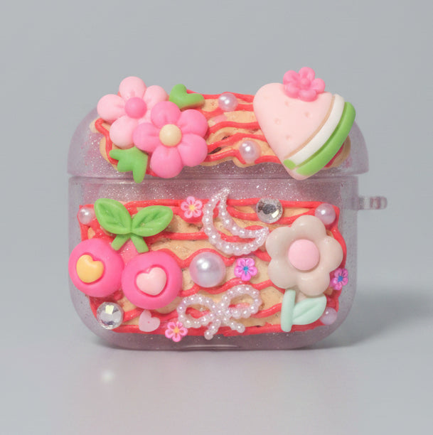 A decorative AirPods case with a colorful floral and heart design, featuring pink flowers, green leaves, a pink macaron, and rhinestone embellishments on a dark background.