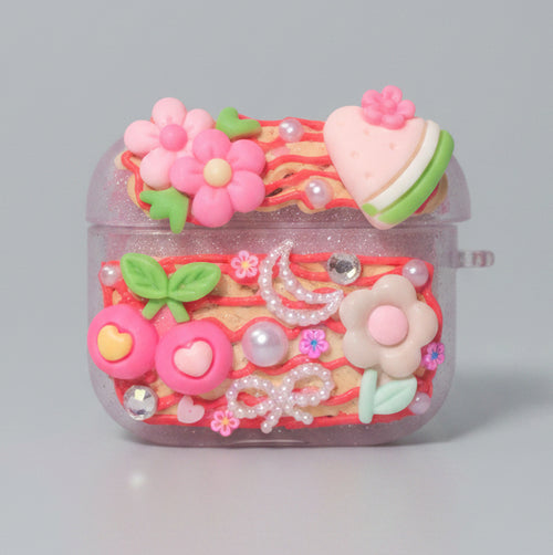 A decorative AirPods case with a colorful floral and heart design, featuring pink flowers, green leaves, a pink macaron, and rhinestone embellishments on a dark background.