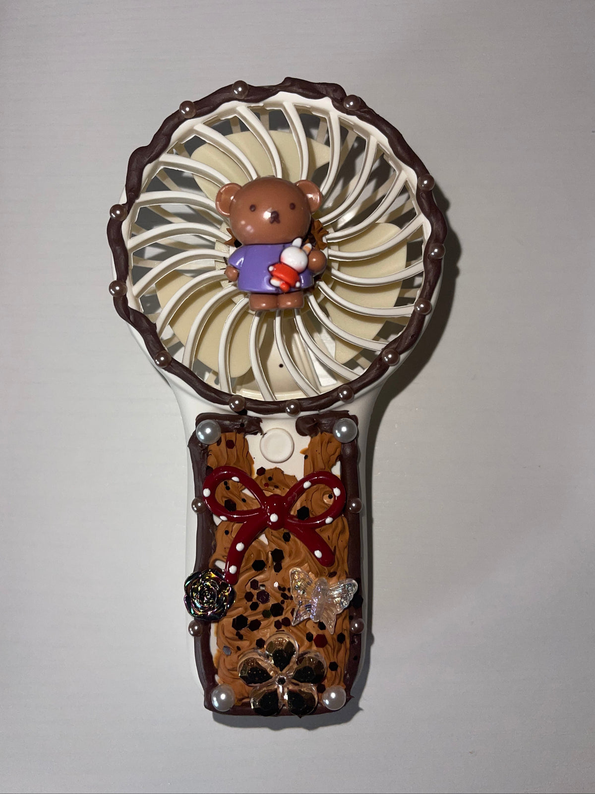 A decorative handheld fan with a brown and beige spotted handle, a red bow detail, and a white ribbed circular head featuring a small brown bear figurine.