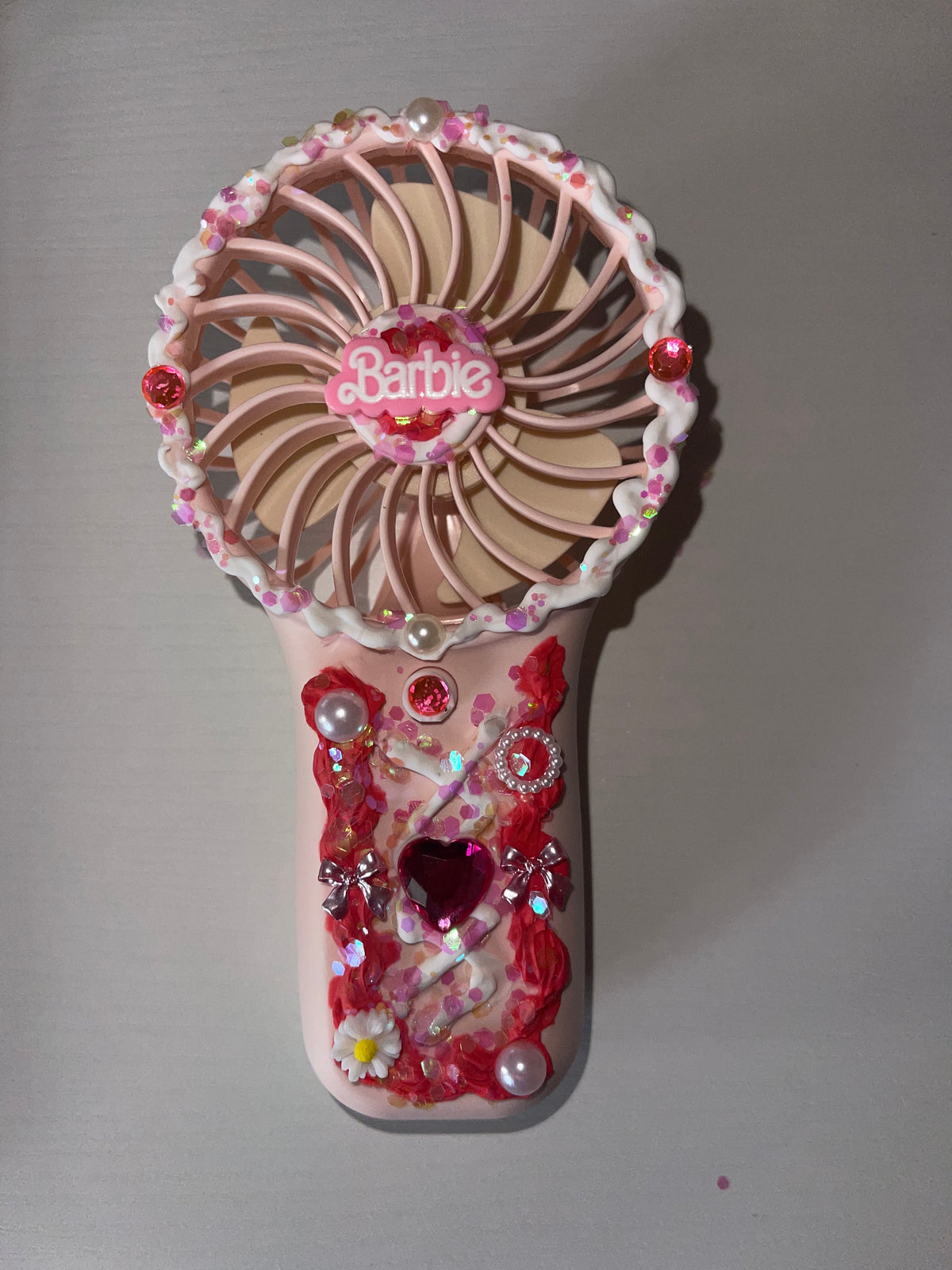 A custom-decorated Barbie-themed handheld fan. The items are decorated with pink and red glitter, faux pearls, rhinestones, and a central pink Barbie logo on the fan.