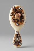 A beige plastic hairbrush decorated with elaborate 3D embellishments, including a brown teddy bear with a pink bow, brown and black glitter, small charms, and rhinestones on the handle.